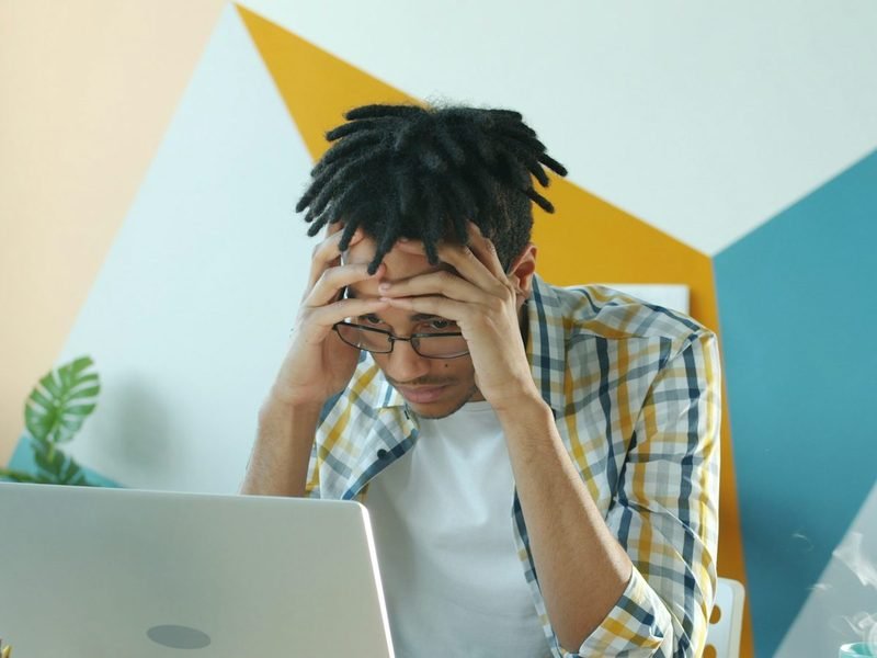 A tired young man holding his head in his hands at a laptop